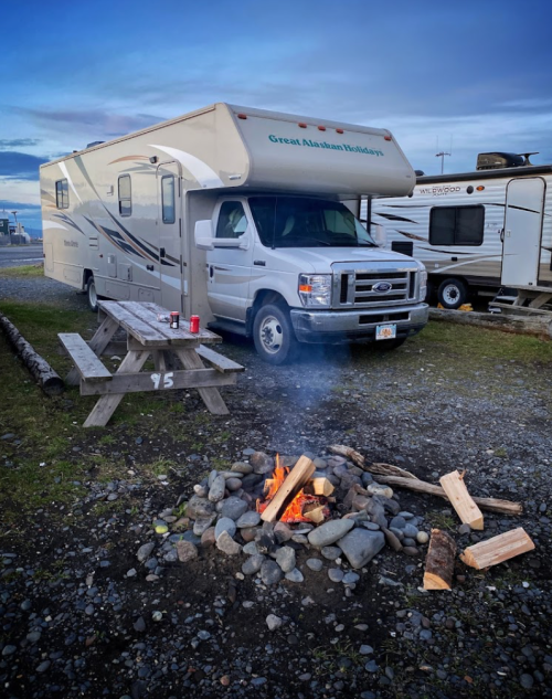 A camper van parked beside a picnic table and a campfire, surrounded by gravel and other RVs in the background.