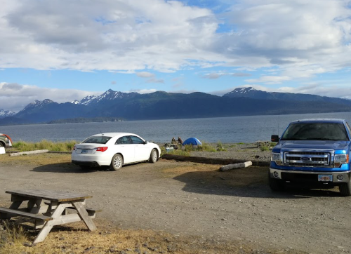 A scenic view of a parking area by the water, with mountains in the background and a few vehicles parked.