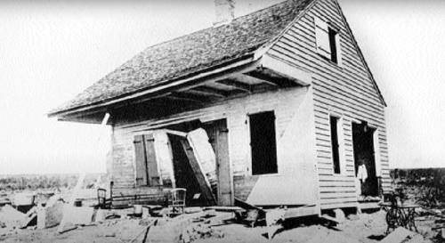 A dilapidated wooden house with a slanted roof and broken windows, surrounded by debris in a barren landscape.