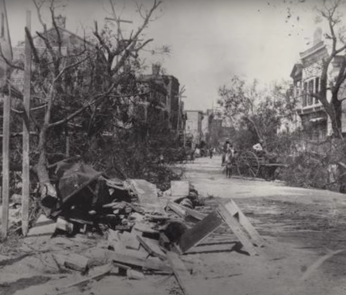 A historic street scene showing debris and fallen trees after a disaster, with buildings and a bicycle in the background.
