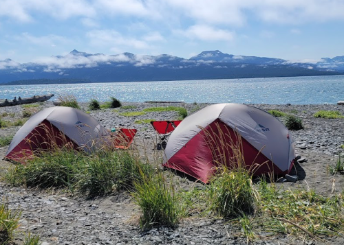 Two red and gray tents on a sandy beach with mountains and a lake in the background under a clear blue sky.