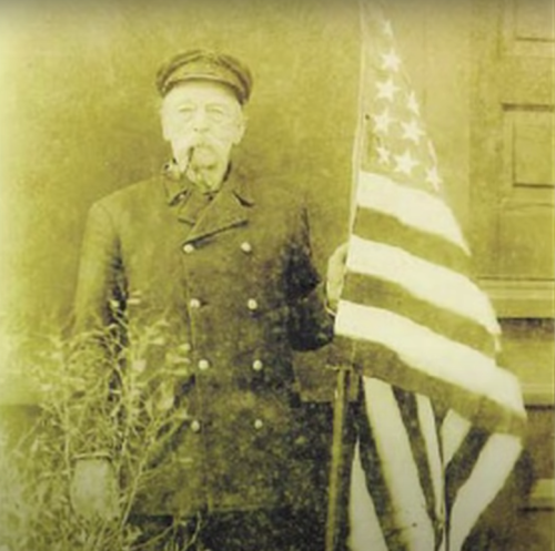 A vintage photo of a man in a coat holding an American flag, with a pipe in his mouth and a serious expression.