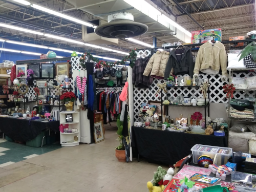 A bustling thrift store aisle filled with clothing, home goods, and decorations, with colorful displays and various items for sale.