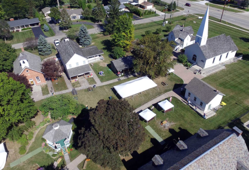 Aerial view of a small community with several buildings, a church, and tents set up on green grass.
