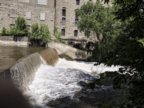 A flowing river cascades over a stone dam, surrounded by greenery and an old stone building in the background.