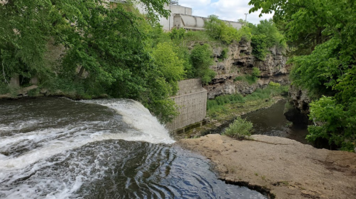 A serene river flows over a rocky edge, surrounded by lush greenery and cliffs in a tranquil natural setting.