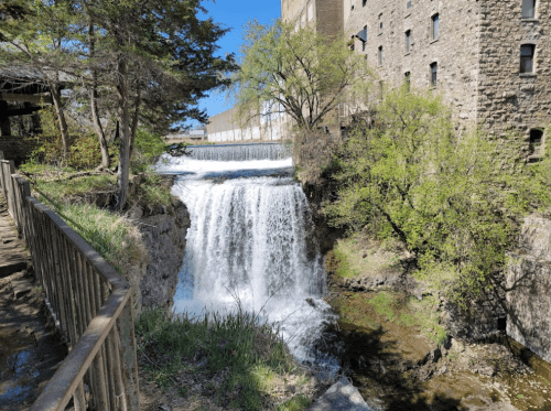 A scenic waterfall cascading over rocks, surrounded by greenery and an old stone building in the background.