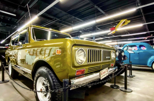 A vintage green SUV displayed in a museum, with bright lights and other classic cars in the background.