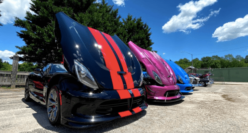 A lineup of three colorful sports cars with hoods raised, parked under a blue sky with fluffy clouds.