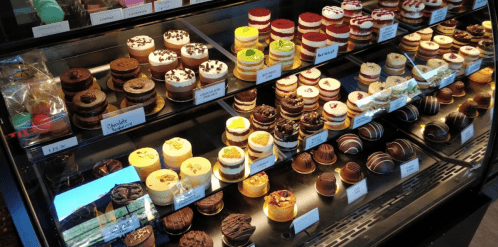 A display case filled with various colorful cakes and desserts, neatly arranged with labels.