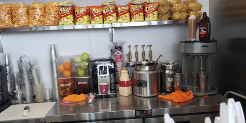 A countertop with snacks, a drink dispenser, fruits, and a blender, set in a food preparation area.