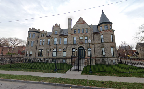 A large, historic stone mansion with turrets and a fenced yard, set against a cloudy sky.