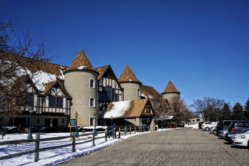 A stone building with turrets and a snowy landscape, alongside a parking lot filled with cars under a clear blue sky.