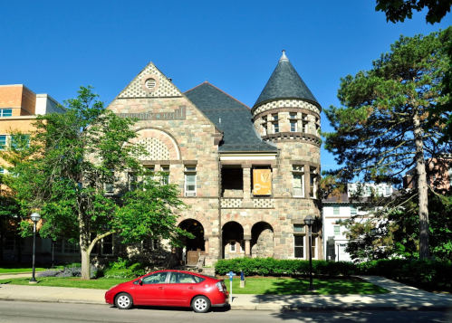 Historic stone building with a turret, surrounded by trees and a red car parked in front, under a clear blue sky.