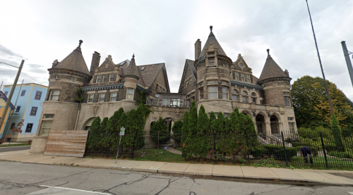 A large, historic mansion with turrets and intricate stonework, surrounded by greenery and a sidewalk.