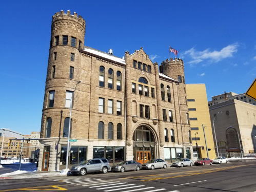 Historic stone building with towers, blue sky, and snow-covered ground, featuring large windows and an American flag.