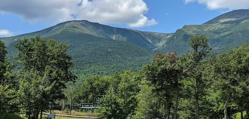 A scenic view of green mountains under a blue sky with fluffy clouds, surrounded by lush trees.