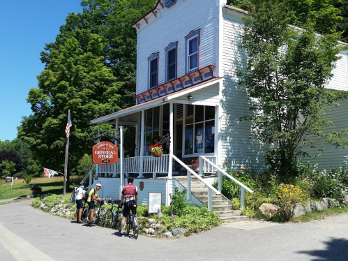 A quaint general store with a porch, surrounded by greenery, as two cyclists pause outside on a sunny day.