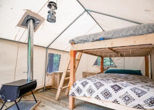 Interior of a cozy tent with a bunk bed, a double bed, and a wood stove, surrounded by wooden flooring.