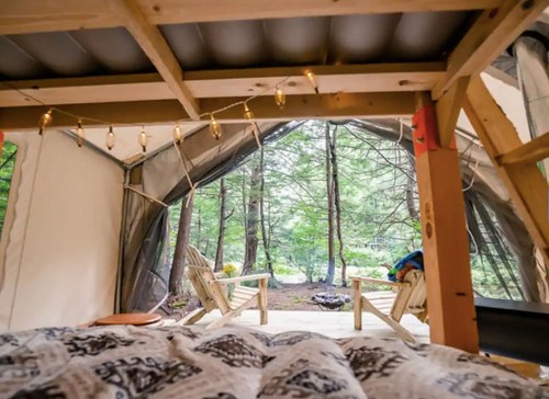 View from inside a cozy tent, featuring a bed and wooden chairs, with trees and a campfire area outside.