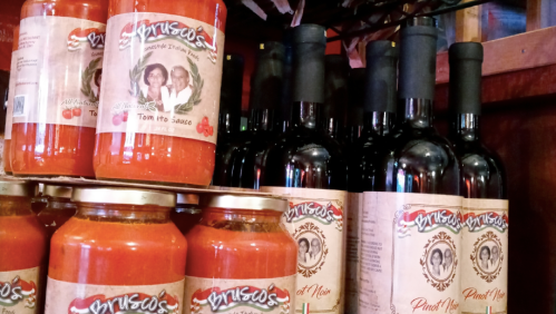 Shelves displaying jars of tomato sauce and bottles of Pinot Noir wine, both labeled with "Brusco's" branding.