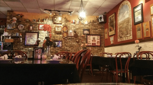 Cozy restaurant interior with brick walls, framed pictures, and tables set with white napkins and flowers.