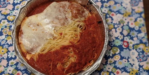 A foil container with spaghetti and marinara sauce topped with melted cheese, placed on a floral-patterned tablecloth.