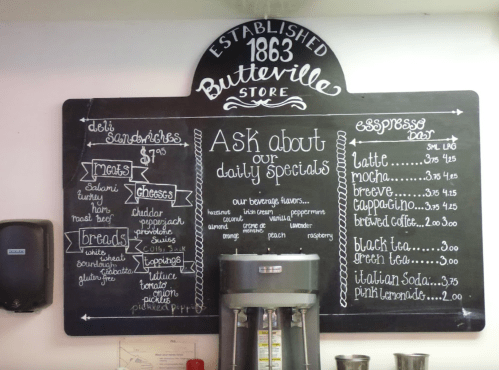 Chalkboard menu at Butterville Store listing deli sandwiches, meats, cheeses, breads, and espresso drinks with prices.