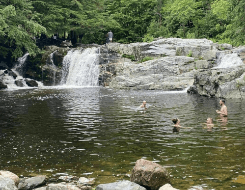 A serene waterfall cascades into a pool where people swim, surrounded by lush greenery and rocky terrain.