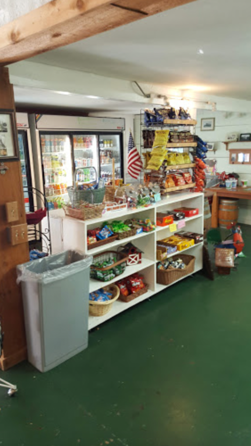 Interior of a small store featuring shelves of snacks, drinks, and a visible American flag. Green flooring and a trash can are present.