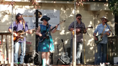 A band performs on stage outdoors, featuring a guitarist, bassist, and two other musicians with instruments.