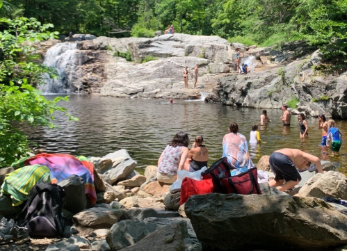 People enjoying a sunny day by a waterfall, swimming and relaxing by a rocky pool surrounded by greenery.