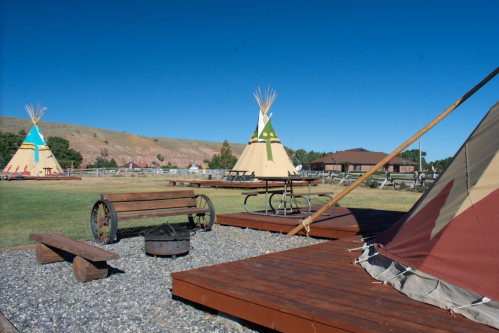 Teepees in a grassy area with picnic tables, a fire pit, and a clear blue sky in the background.