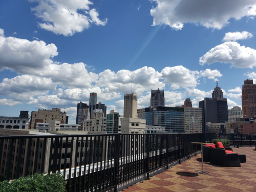 A rooftop view of a city skyline with tall buildings and a bright blue sky dotted with fluffy clouds.