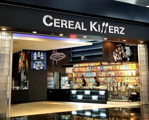 A colorful cereal bar named "Cereal Killerz" with various cereal boxes displayed and staff serving customers.