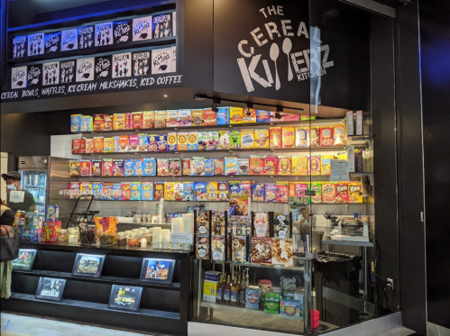 A colorful cereal shop display featuring various cereal boxes, milk, and toppings, with a modern black storefront.