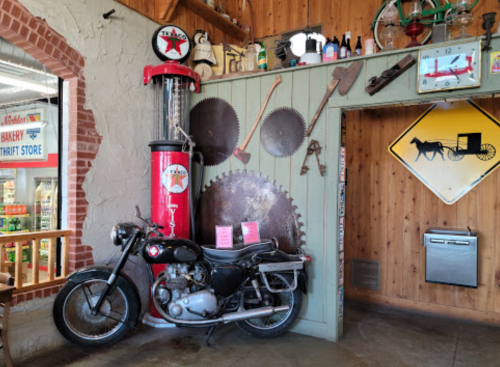 A vintage motorcycle next to a retro gas pump and various tools on a wooden wall in a rustic setting.