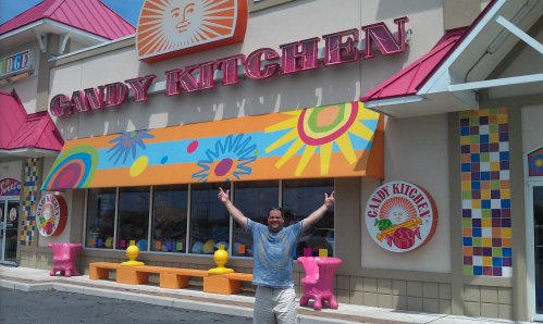 A man stands outside the colorful Candy Kitchen store, smiling and posing with his arms raised in excitement.