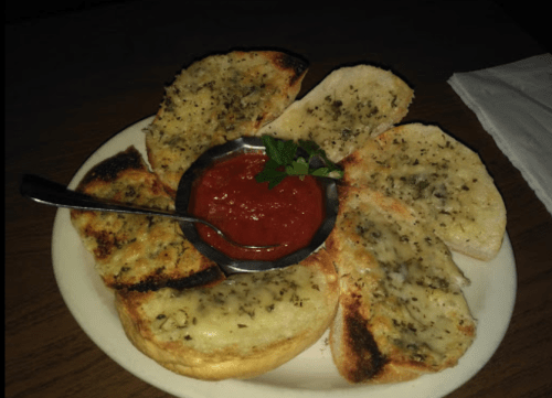 A plate of toasted bread slices topped with herbs, served with a small bowl of marinara sauce and a sprig of parsley.