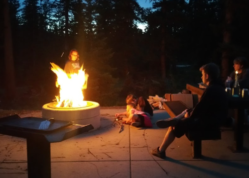 A group of people gathered around a large campfire in a forested area at dusk, enjoying the warmth and ambiance.