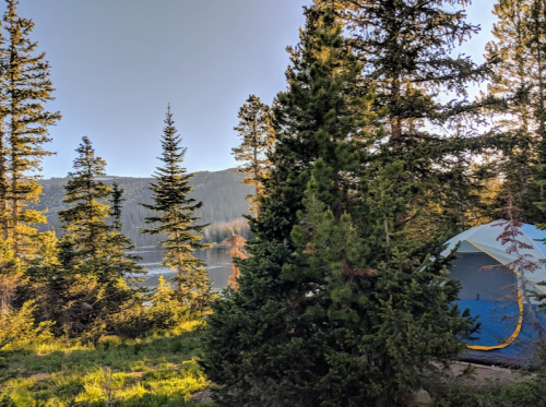 A serene campsite by a lake, surrounded by tall trees and mountains under a clear blue sky.