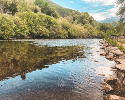 A serene river flows through lush greenery, with smooth stones lining the bank under a clear blue sky.