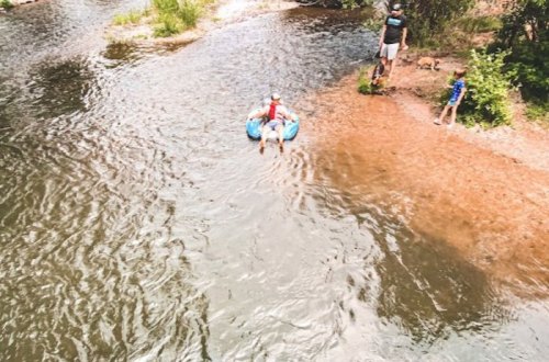 A child floats on a blue inflatable in a river, while two people and a dog stand on the shore nearby.