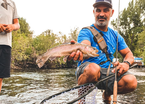 A man kneels by the water, holding a rainbow trout, with a fishing net nearby and a person standing in the background.