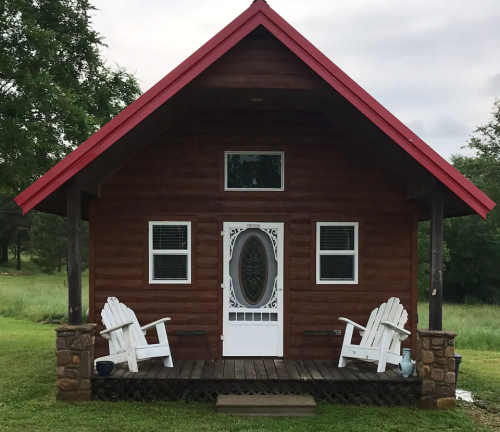 A small wooden cabin with a red roof, white door, and two white chairs on a porch, surrounded by greenery.