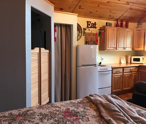 Cozy kitchen with wooden cabinets, a refrigerator, and a stove, viewed from a bedroom with a bed and decorative elements.