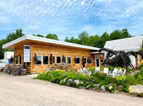 A rustic wooden building with large windows, surrounded by greenery and outdoor seating, under a blue sky.