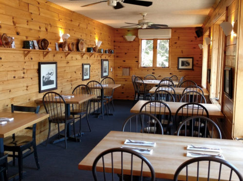 Cozy wooden restaurant interior with tables, black chairs, and framed photos on the walls. Natural light from windows.
