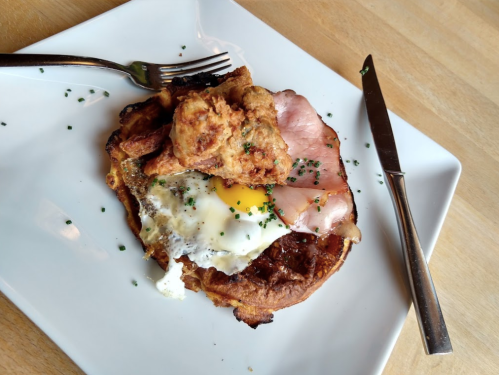 A plate with a crispy fried chicken piece, ham, a sunny-side-up egg, and a savory pancake, garnished with chives.