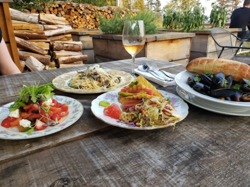 A rustic table displays a salad, pasta, mussels, and a loaf of bread, with a glass of wine in a scenic outdoor setting.